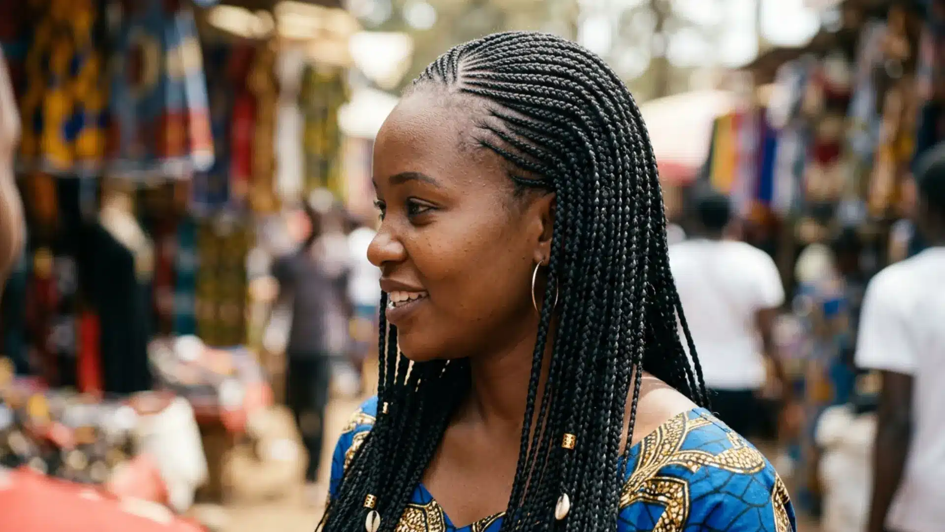 Side profile of a smiling Black woman with neat Fulani braids, hair beads, and cowrie shells at an outdoor market