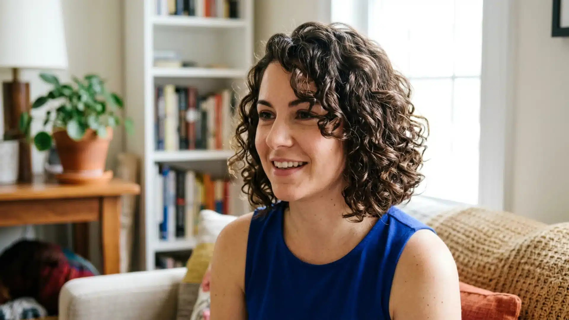 Smiling woman with short, curly brown hair wearing a blue top sits in a living room