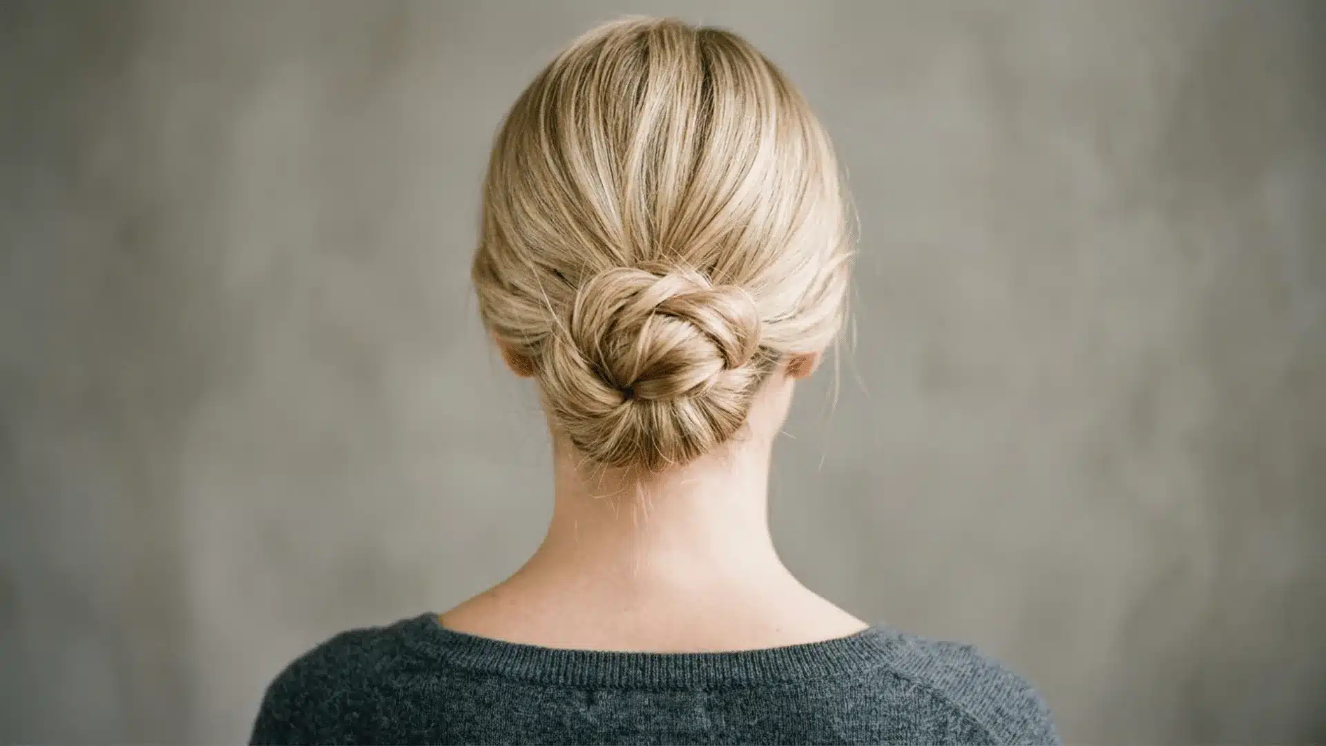 The back of a blonde woman's head showing a neatly styled, twisted low bun hairstyle against a grey wall