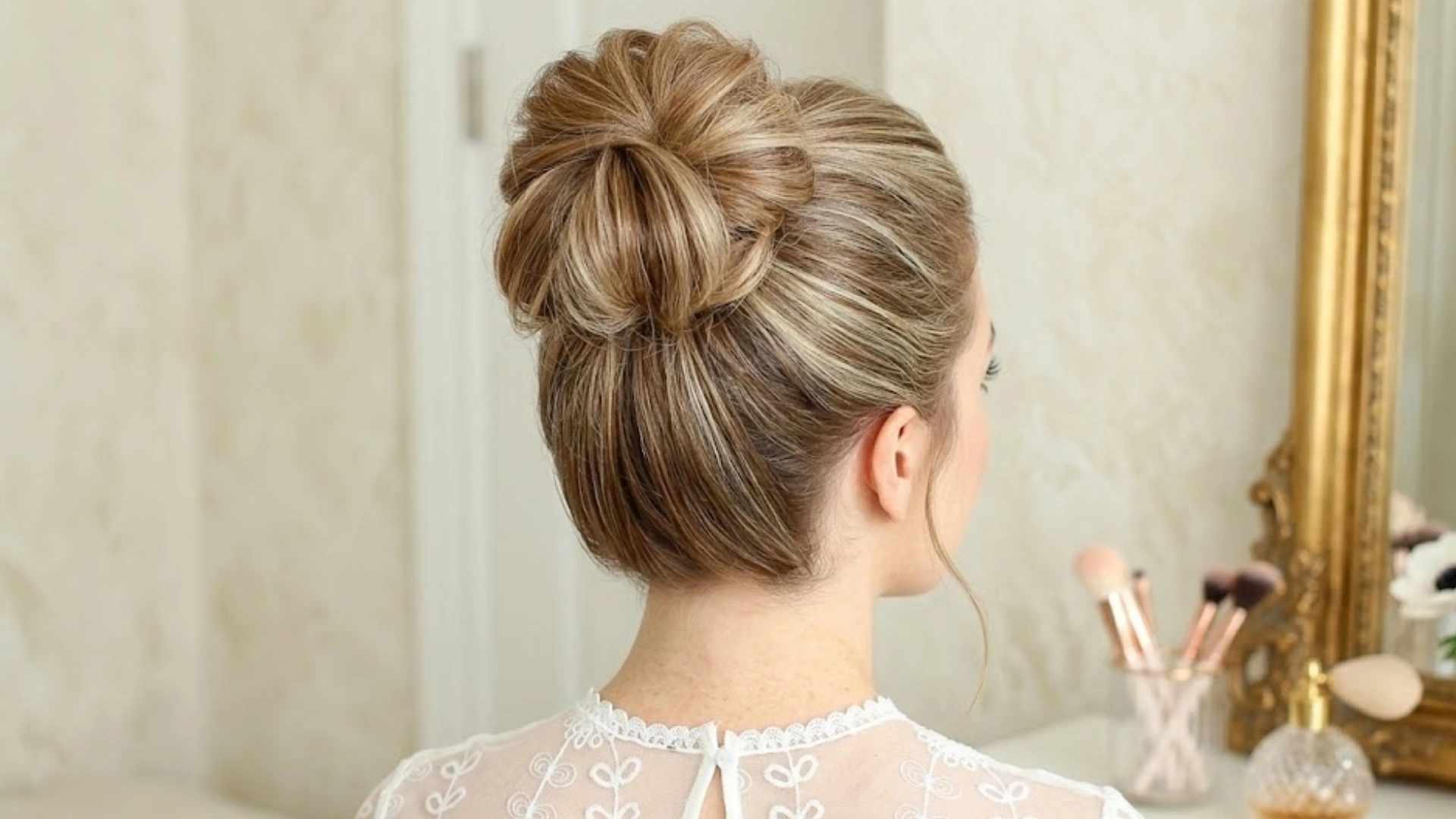 Woman with a voluminous messy bun hairstyle viewed from behind while sitting at a vanity mirror indoors