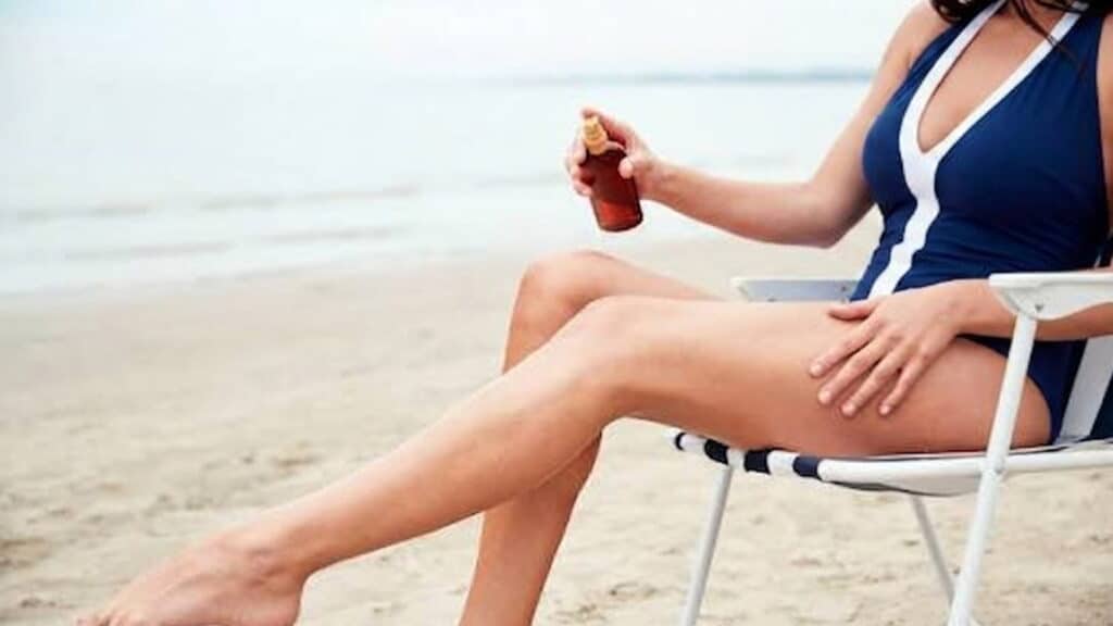 Woman with tan legs in a navy one-piece swimsuit applies tanning oil from a bottle while sitting in a beach chair