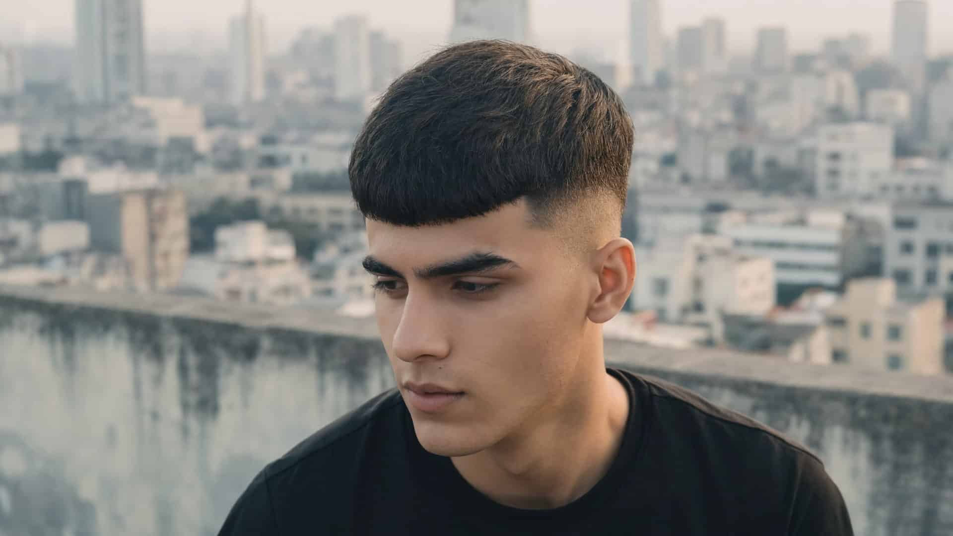Young man with a textured French crop haircut looking down on a rooftop with a city background