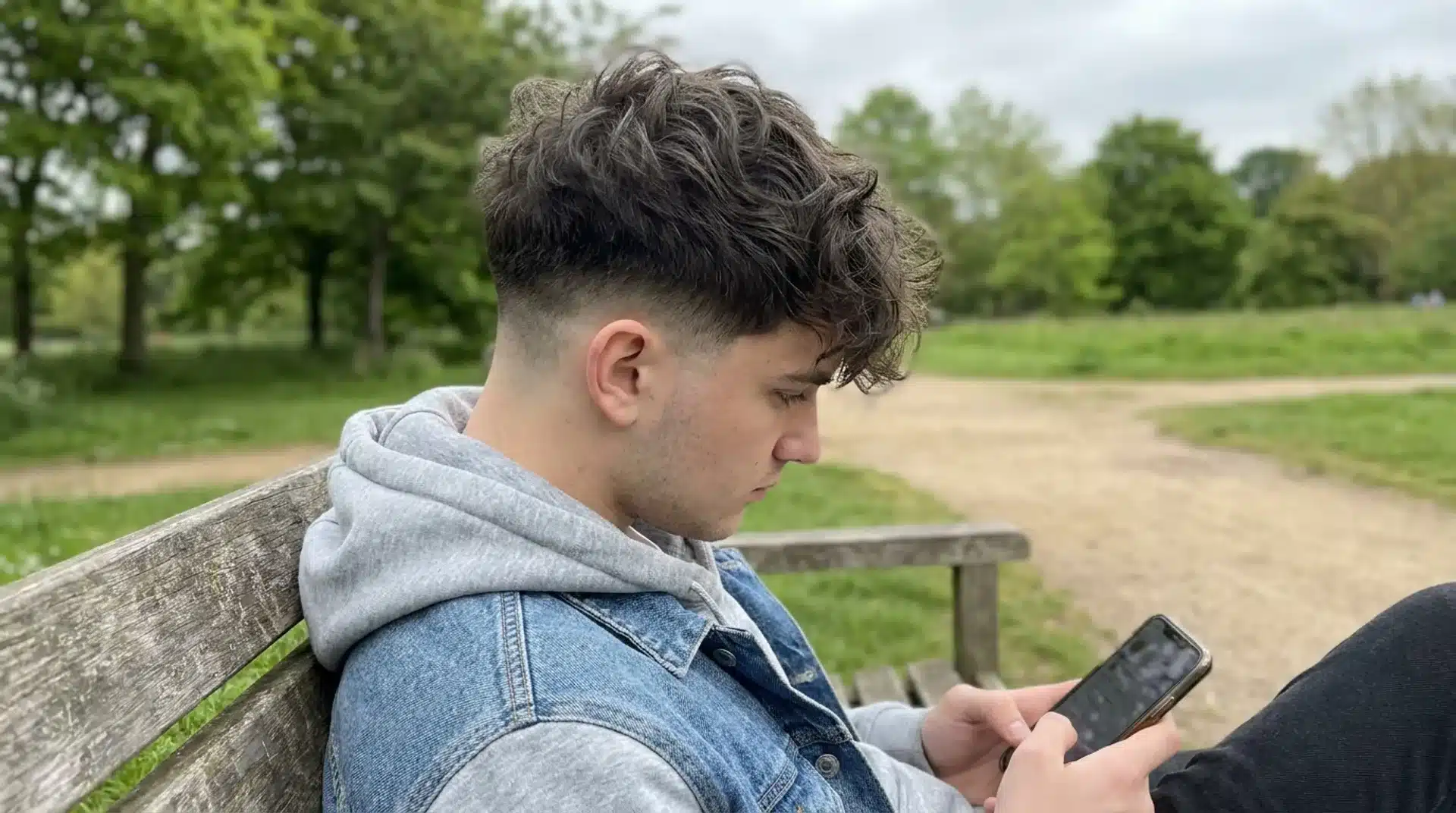 Young man with messy textured low taper fade using a phone on a park bench with trees behind him