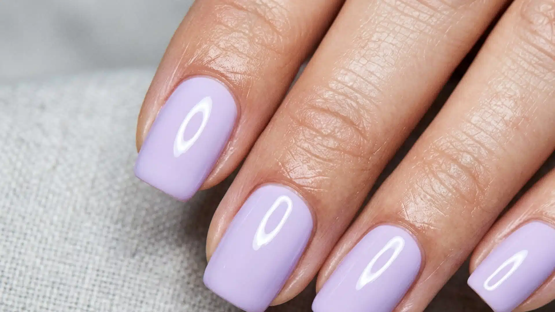 A hand with glossy, lavender-colored square nails resting on a textured fabric