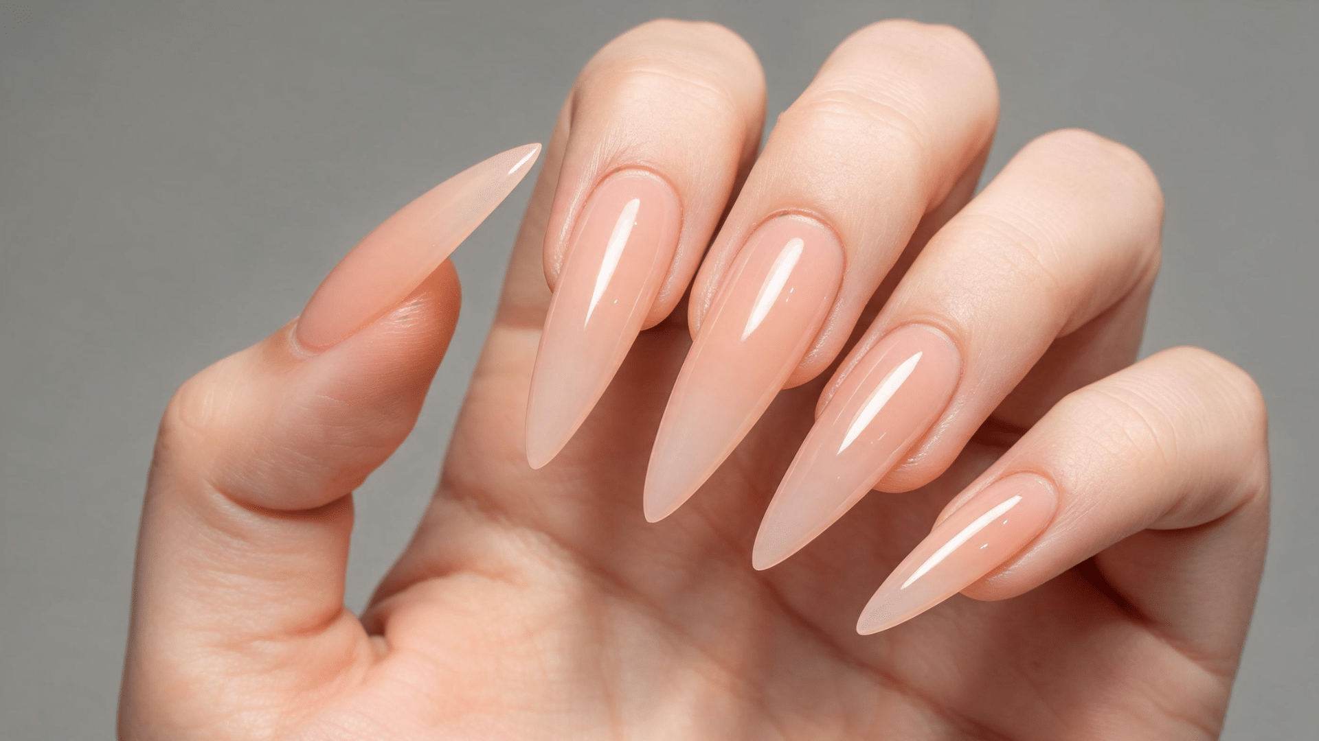 a hand with long, sharp stiletto nails in a translucent, glossy peach shade against a gray background