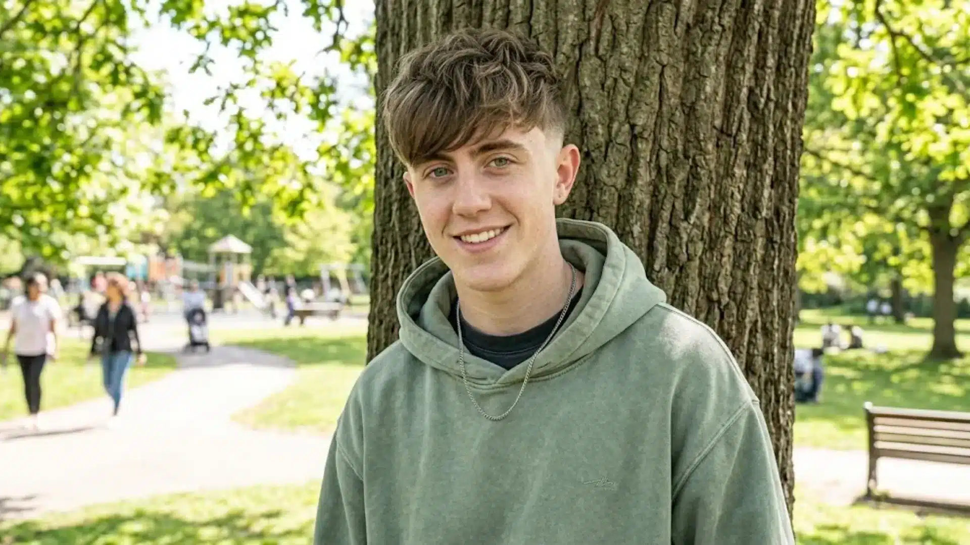 A smiling man in a green hoodie stands against a tree in a sunny park with a playground in the background
