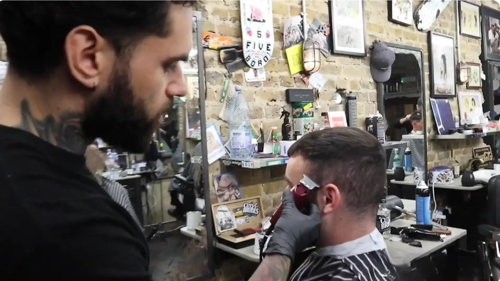 Barber trimming client hair with clipper in busy barbershop, brick wall, mirror, tools and grooming products