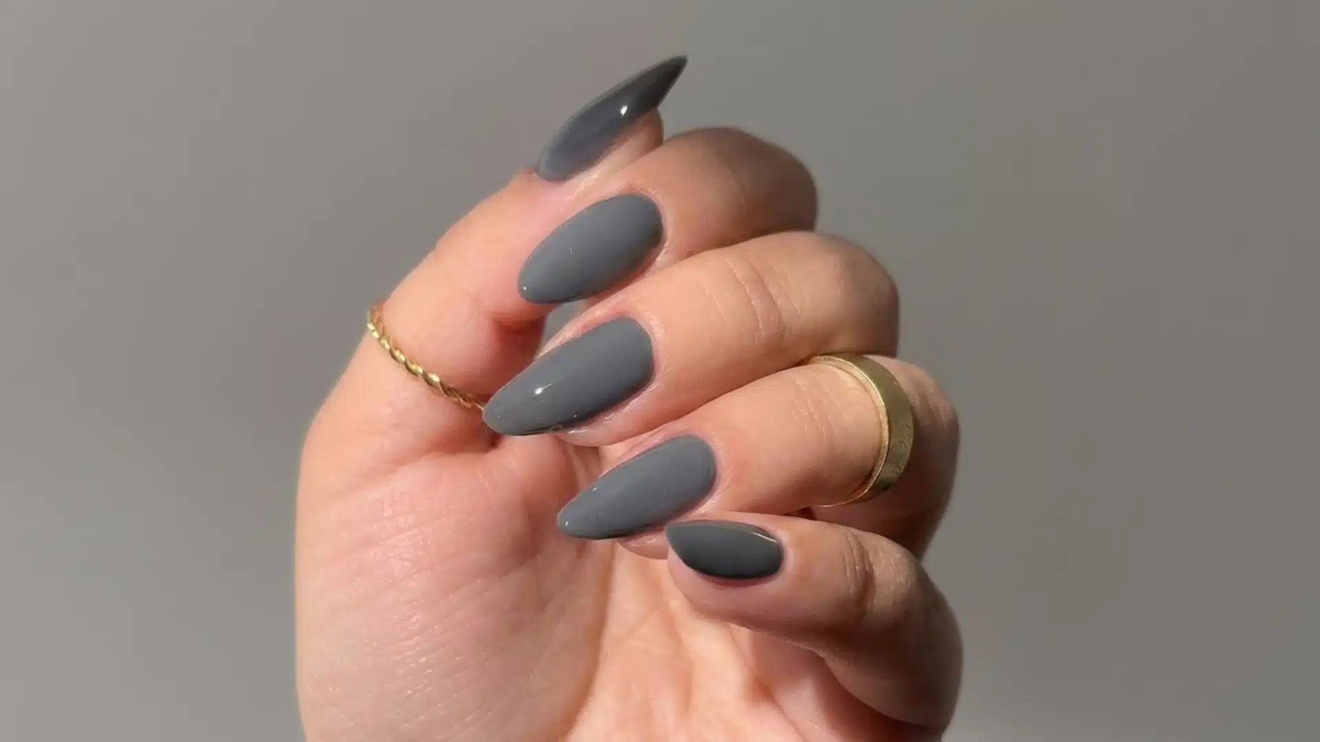 charcoal gray almond nails with a glossy finish and gold rings on a natural hand against a soft gray background