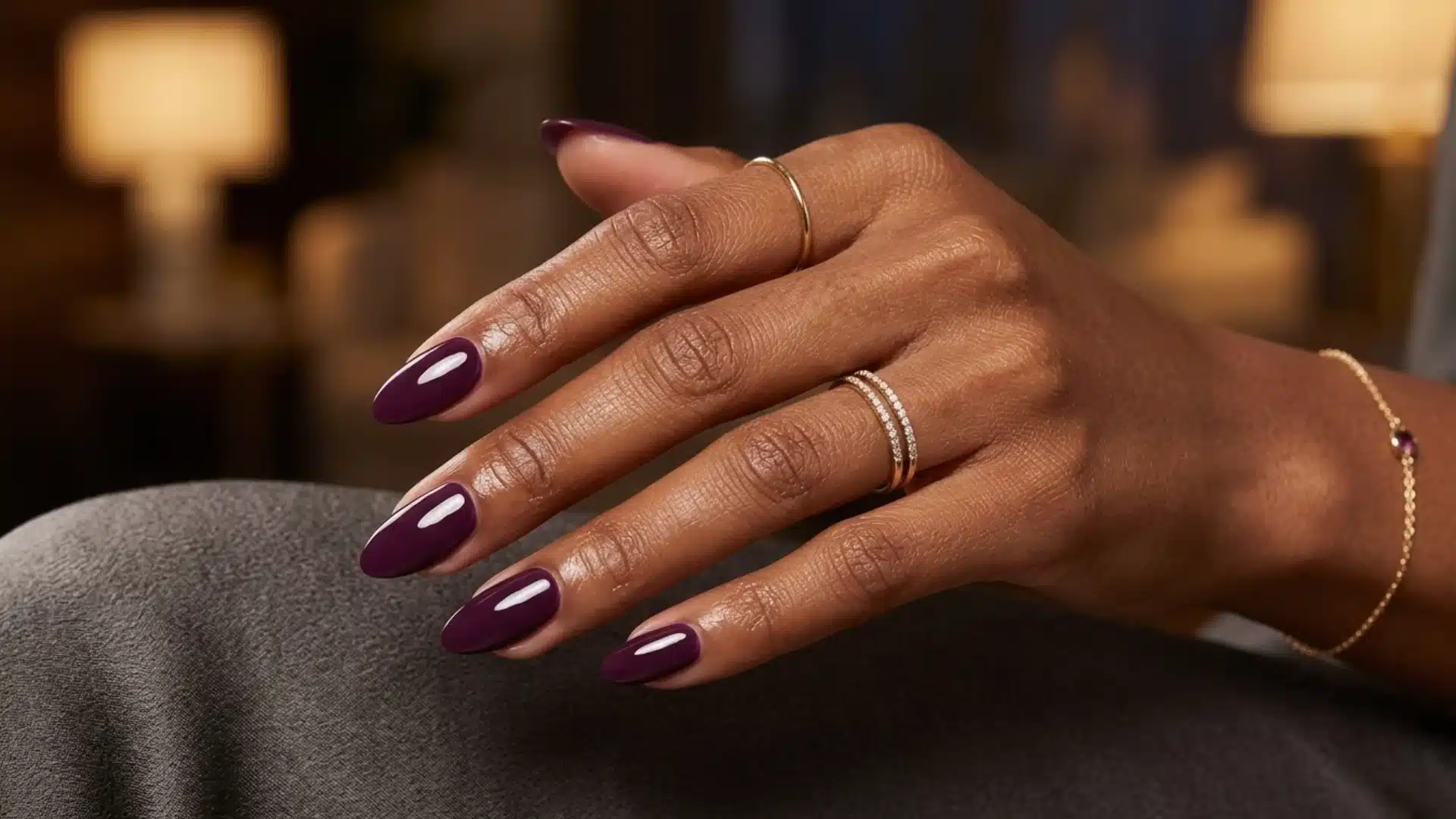 dark plum almond nails with glossy finish on hand wearing gold rings in warm indoor lighting
