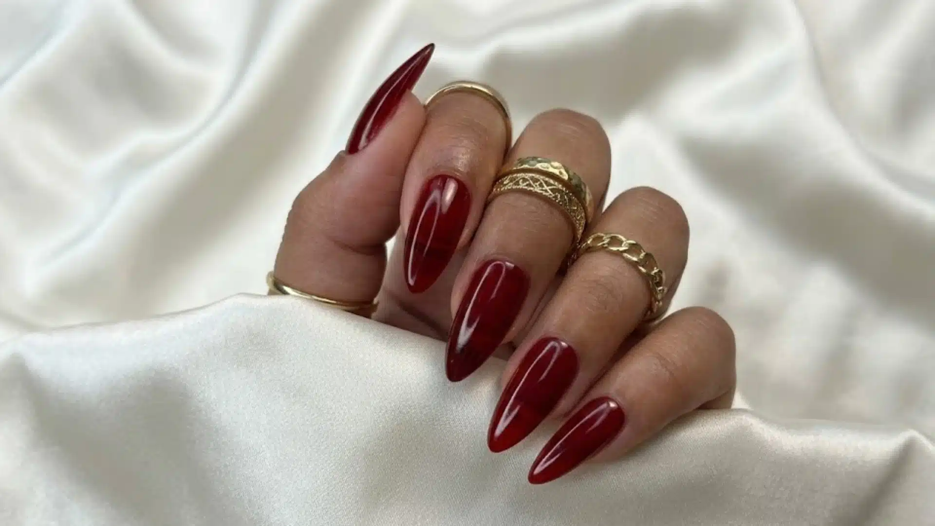 Deep red glossy stiletto nails with gold rings on hand resting on white satin fabric backdrop