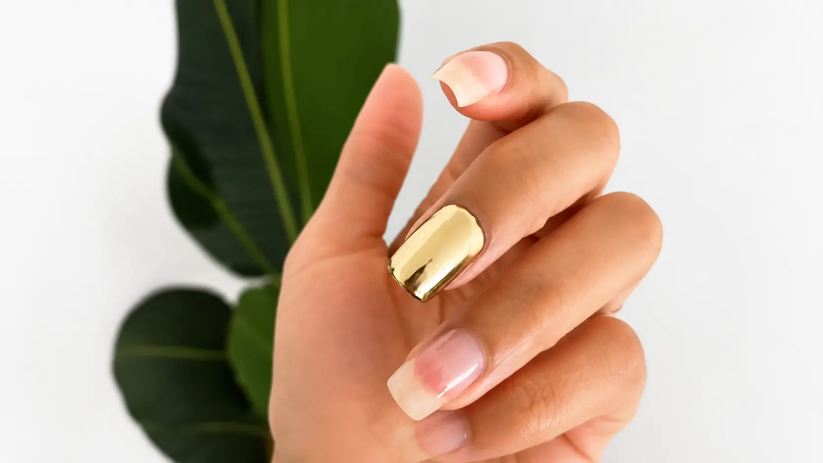 gold chrome nail design on a hand with glossy natural nails against a clean white background and green leaf backdrop