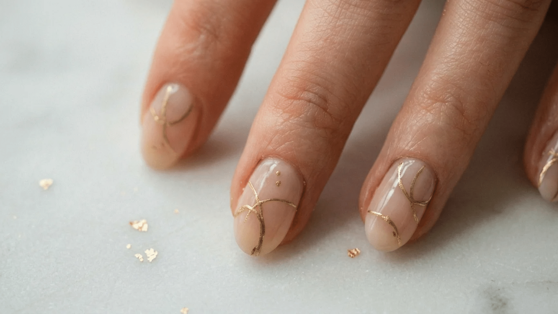 gold line art almond nails with nude base polish and delicate metallic details on fingers over white surface