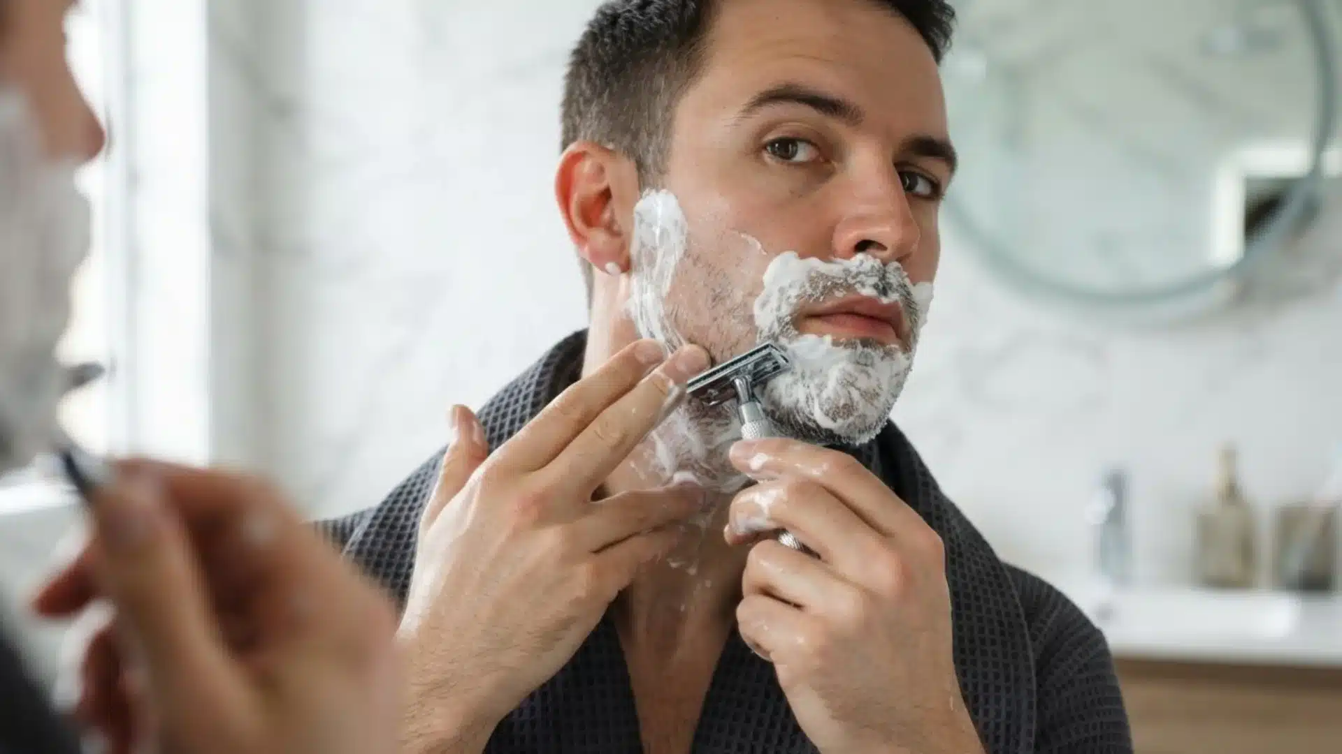 Man shaving with a razor and shaving cream while looking into a bathroom mirror