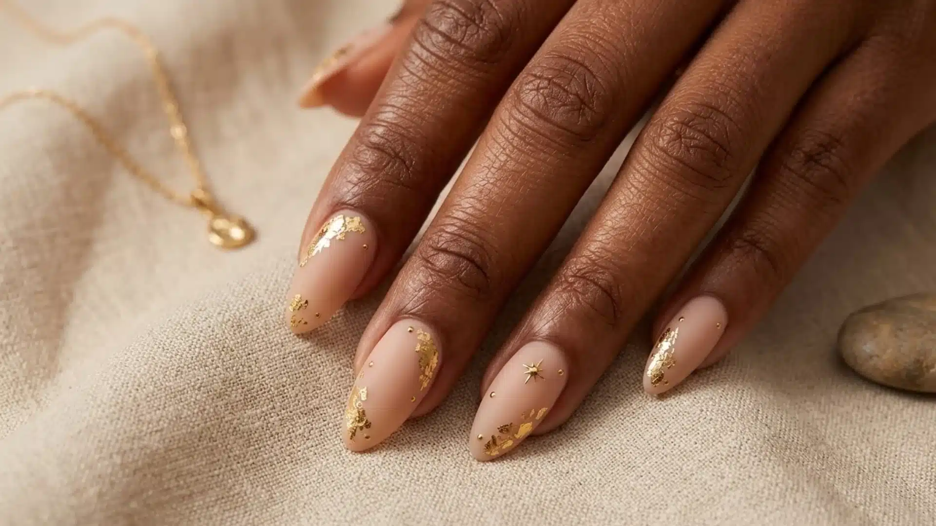 matte nude almond nails with gold foil accents and delicate details on dark skin hand over fabric surface