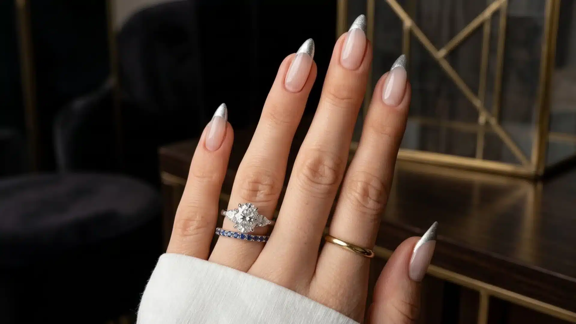 metallic silver french tip almond nails with nude base polish on hand wearing rings in elegant indoor setting
