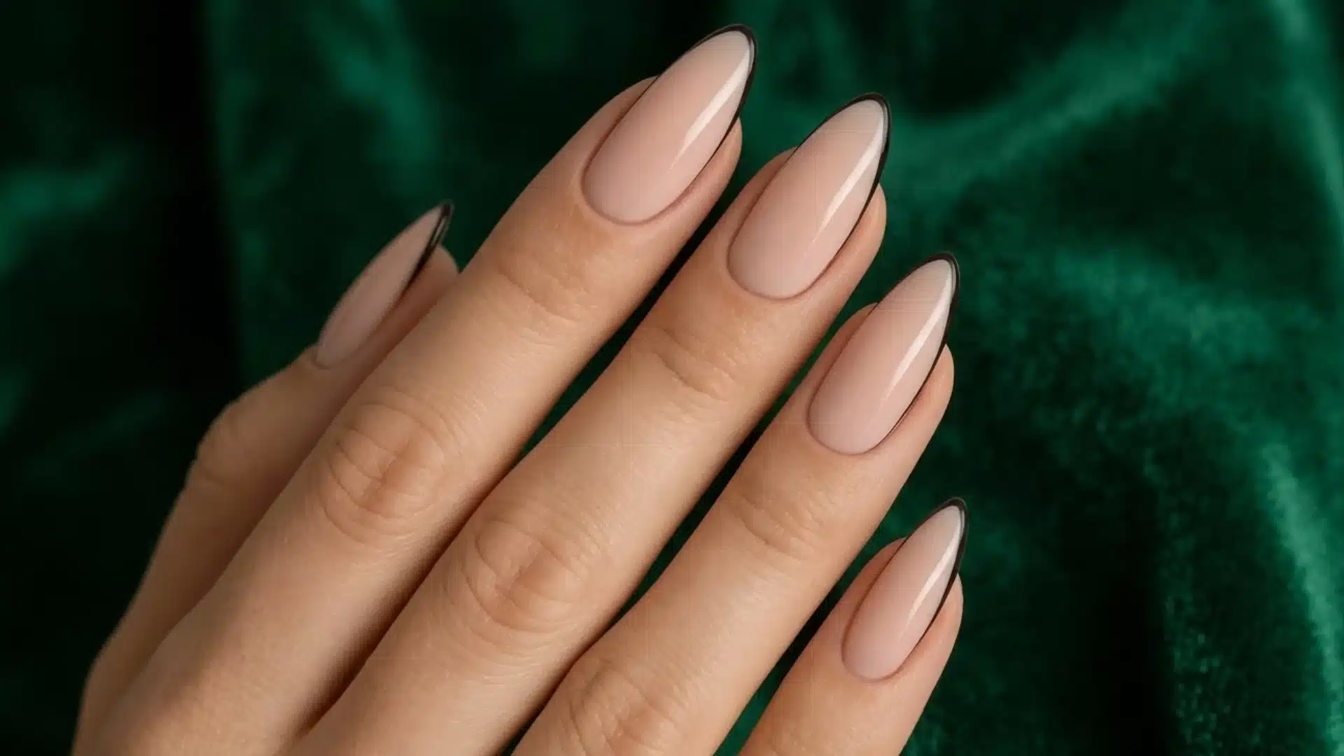 nude almond nails with thin black french tips and glossy finish on hand against dark green background