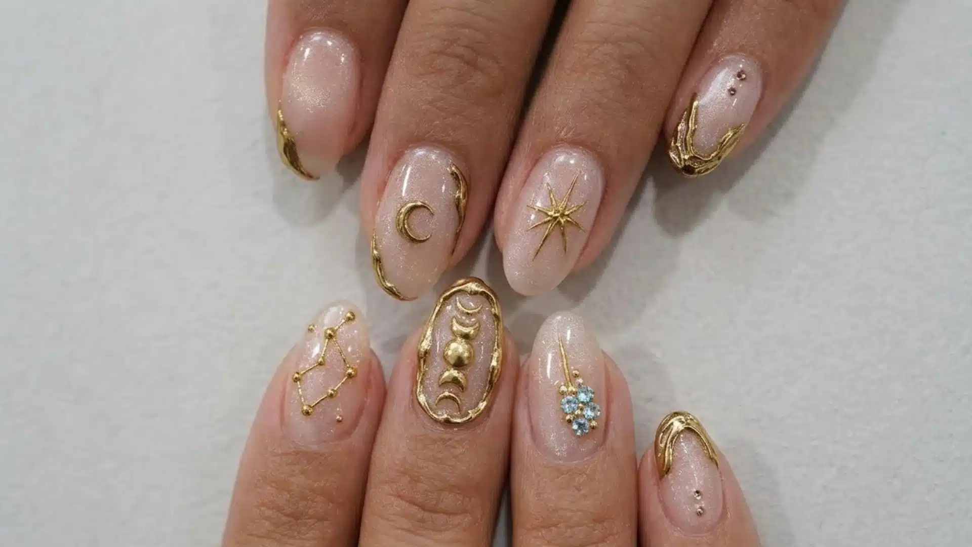 nude negative space almond nails with gold celestial art and delicate metallic details on fingers over white surface