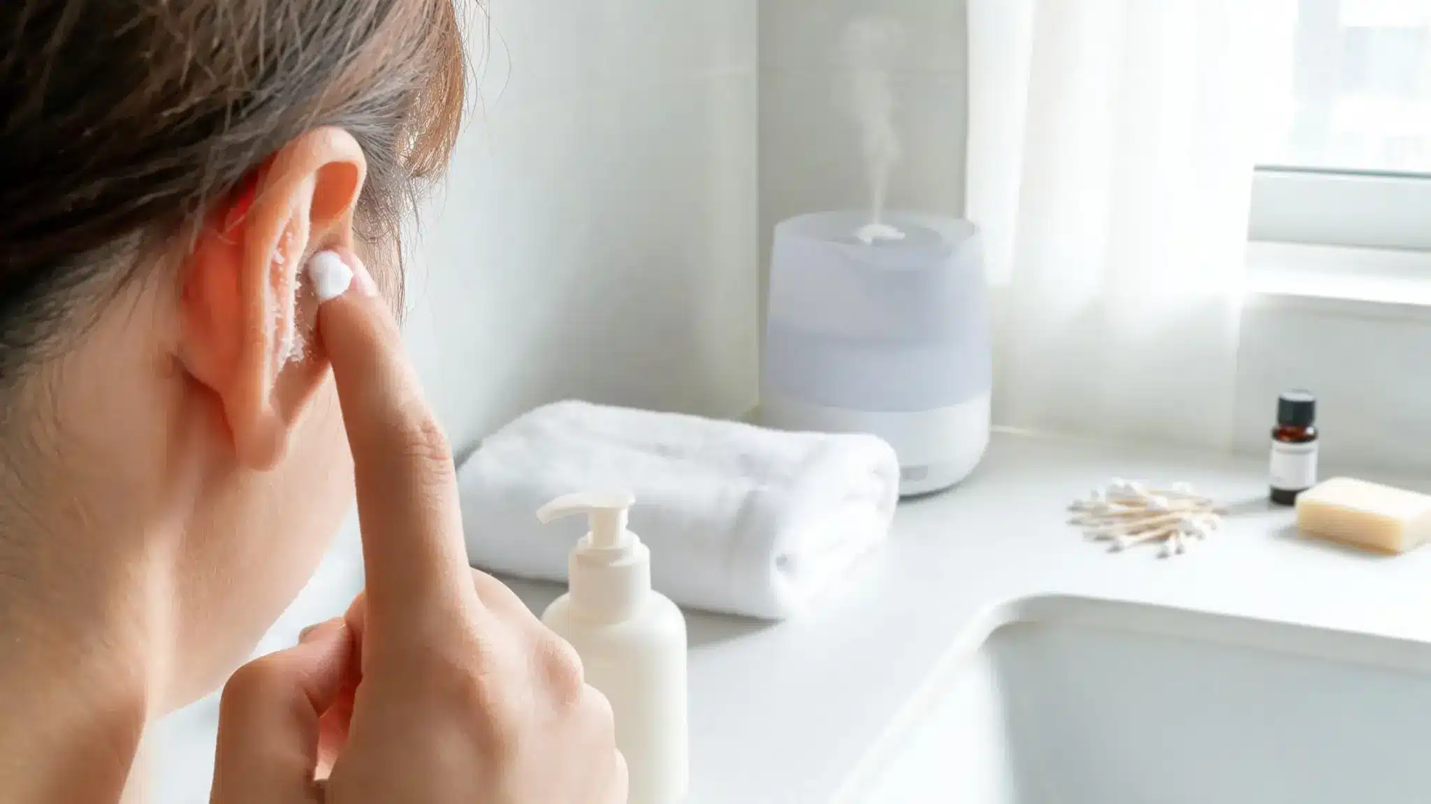 person applying moisturizer to dry flaky outer ear with gentle skincare items in bathroom setting