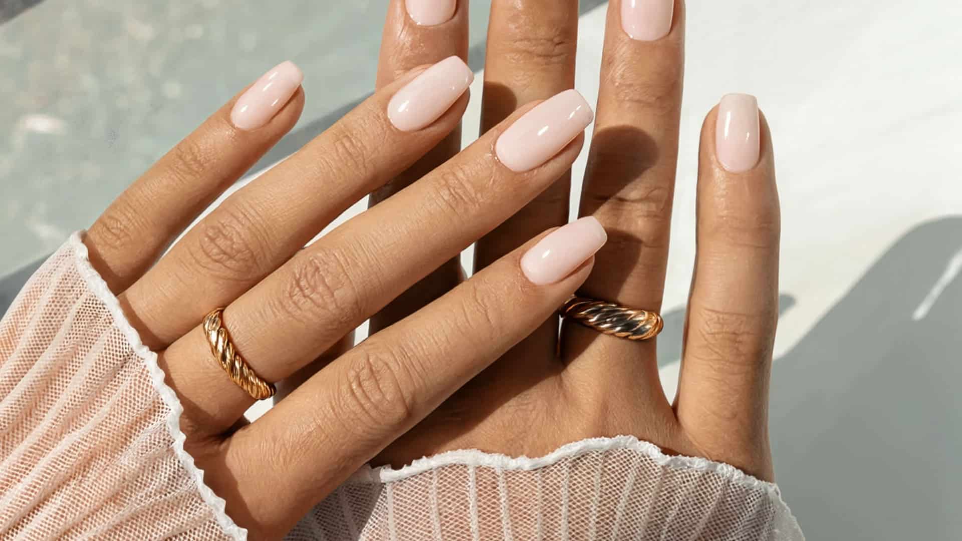 shows two hands with glossy, pale pink square nails and gold rings, set against a bright backdrop
