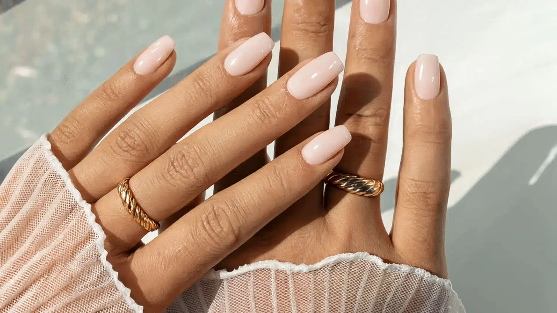 shows two hands with glossy, pale pink square nails and gold rings, set against a bright backdrop