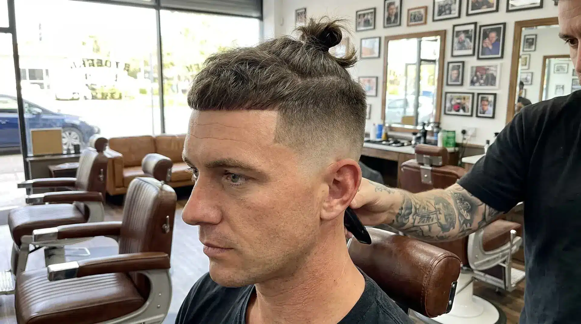 Side profile of a man in a barbershop getting a high fade haircut with a textured top knot and blunt fringe.