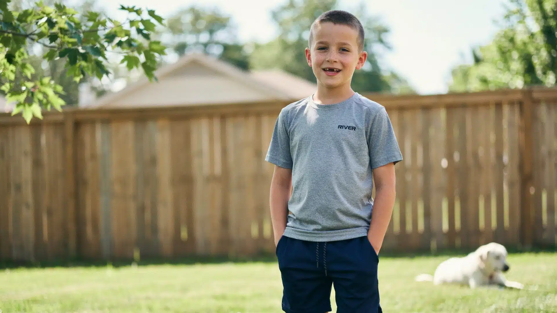 smiling boy with a buzz cut fade wearing a gray shirt standing in a sunny backyard with a dog nearby
