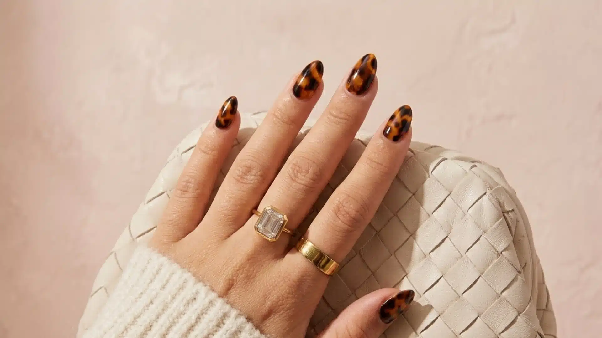 tortoiseshell almond nails with brown and black spotted pattern on hand wearing gold rings over woven background
