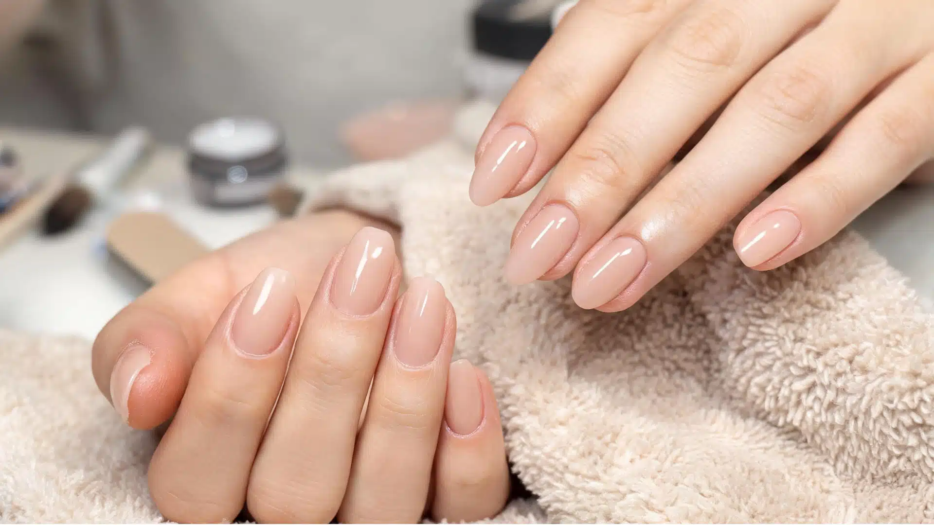 Two hands with glossy, nude oval nails resting on a fluffy beige towel at a salon station
