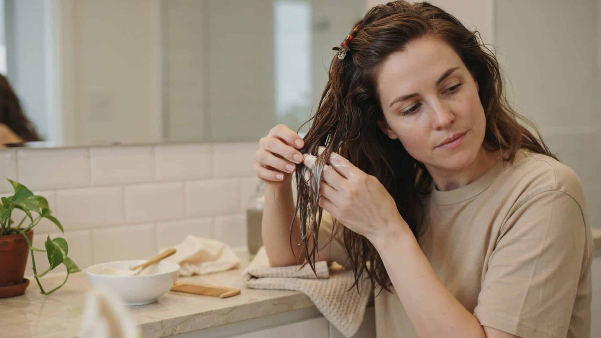 woman applying a creamy mayonnaise hair mask through damp wavy hair beside a bowl and wide tooth comb