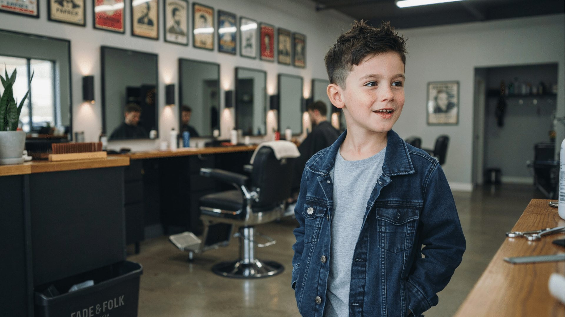 young boy with a spiky fade haircut wearing a denim jacket and smiling inside a modern barbershop