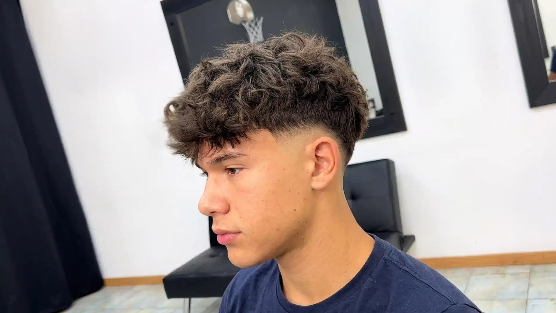 young man with a curly fringe fade haircut and textured brown hair seated inside a modern barbershop