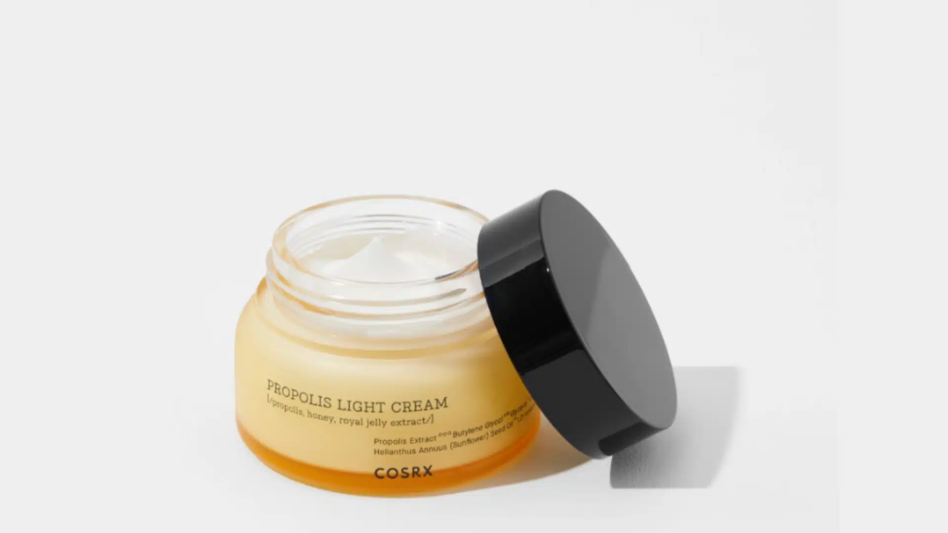 cosrx propolis light cream jar open with black lid resting beside it on a white background