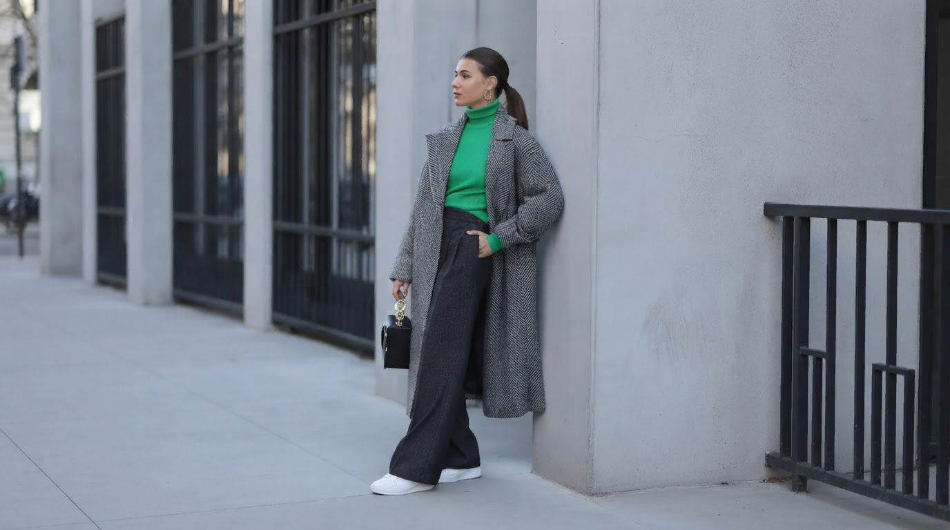 woman in dark grey trousers with a bright green knit and long grey coat on a city sidewalk